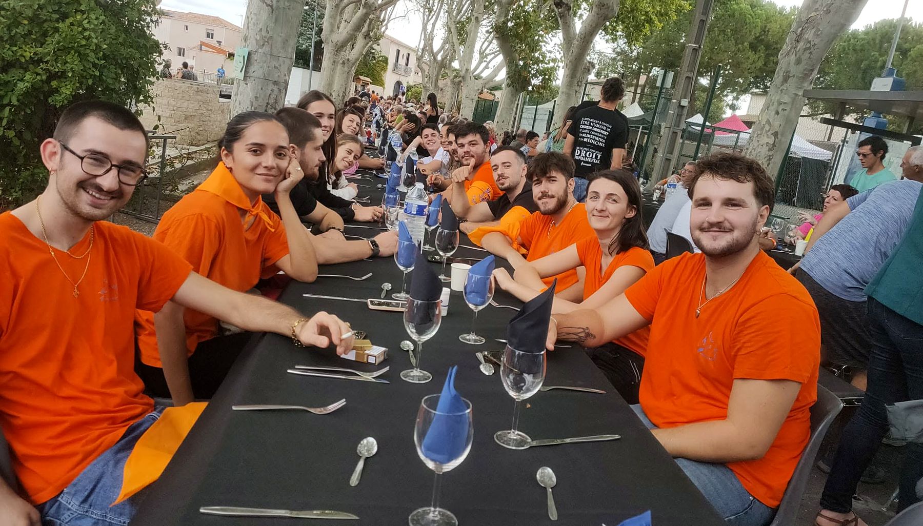 Longue tablée dressée sous les platanes de Aumes pour le repas de la fête du village, bénévoles en t-shirts orange.